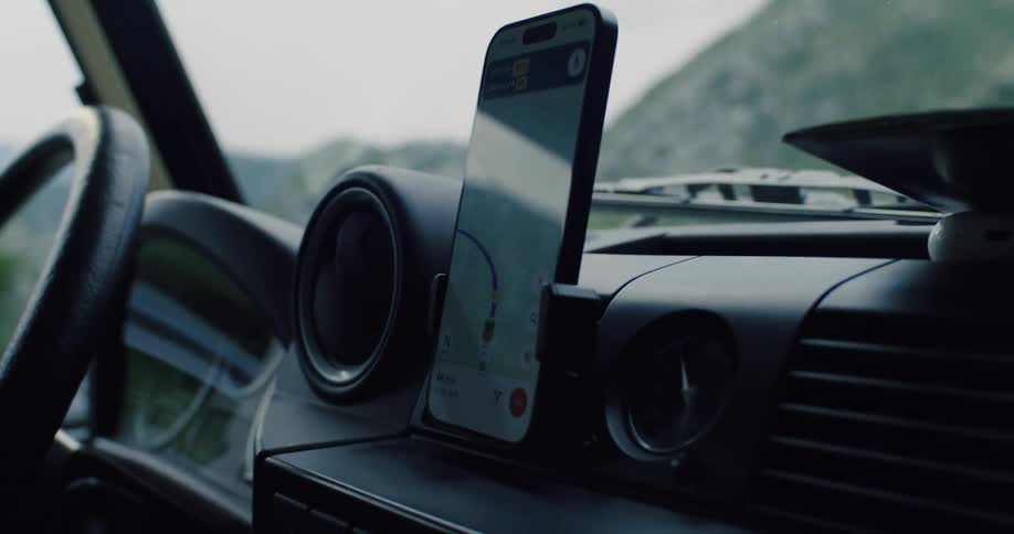 A close-up view from inside a car shows a smartphone on the dashboard using a GPS map app to navigate a winding road through the mountains