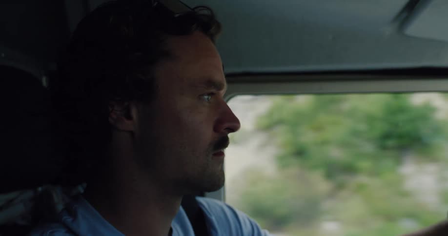A pensive man with a mustache drives a car on a road trip, seen in a close-up profile