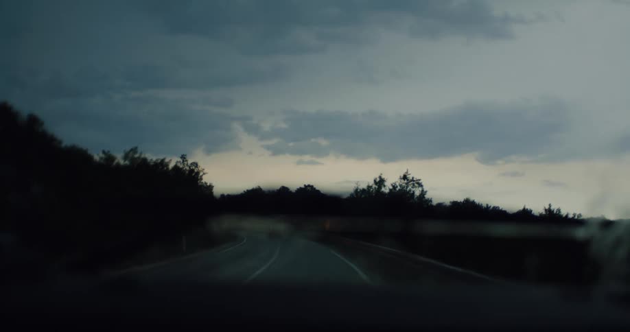 A moody point-of-view shot from a car driving on a wet, rural road at dusk, with windshield wipers clearing away the rain