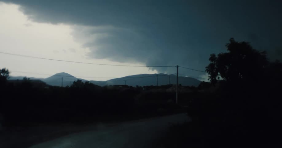 A POV shot from inside a car driving on a rural road as a dramatic, dark storm gathers over the mountains in the distance