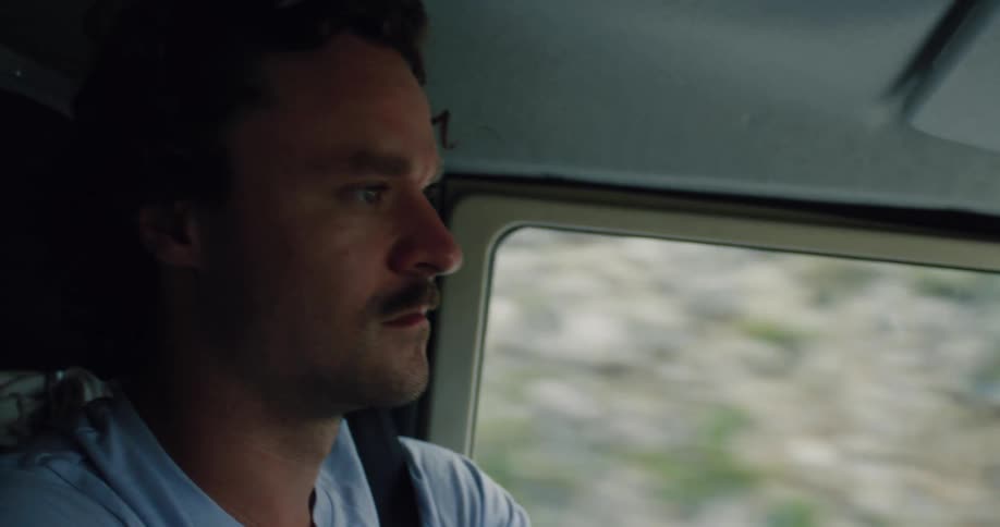 A man with a mustache and tattoo drives a vintage 4x4 on a rugged road trip, with a focus on his hand on the wheel and his determined expression