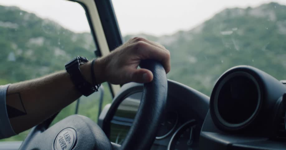 Close-up of a man's tattooed hand gripping the steering wheel while driving a classic off-road vehicle through a mountainous landscape