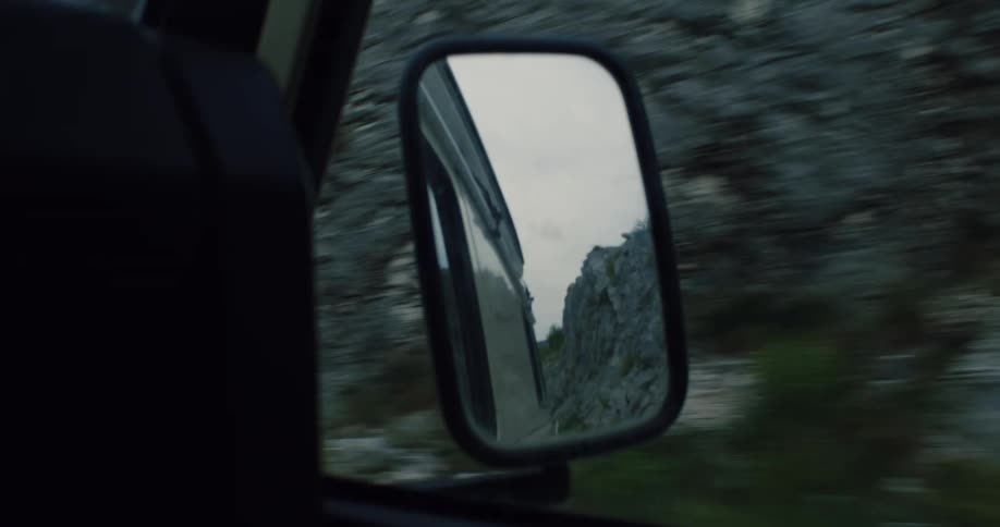 A rugged mountain landscape is reflected in the side mirror of a moving car on a scenic road trip