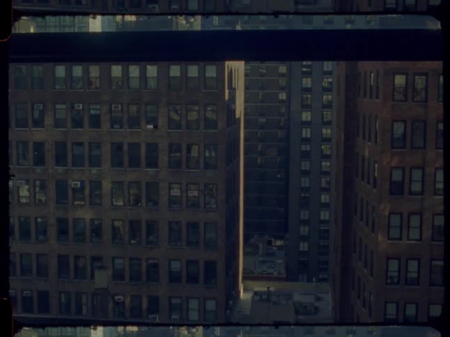 A vintage film shot slowly pulls back from a sunlit alley between tall city buildings to reveal the view from a dark room window