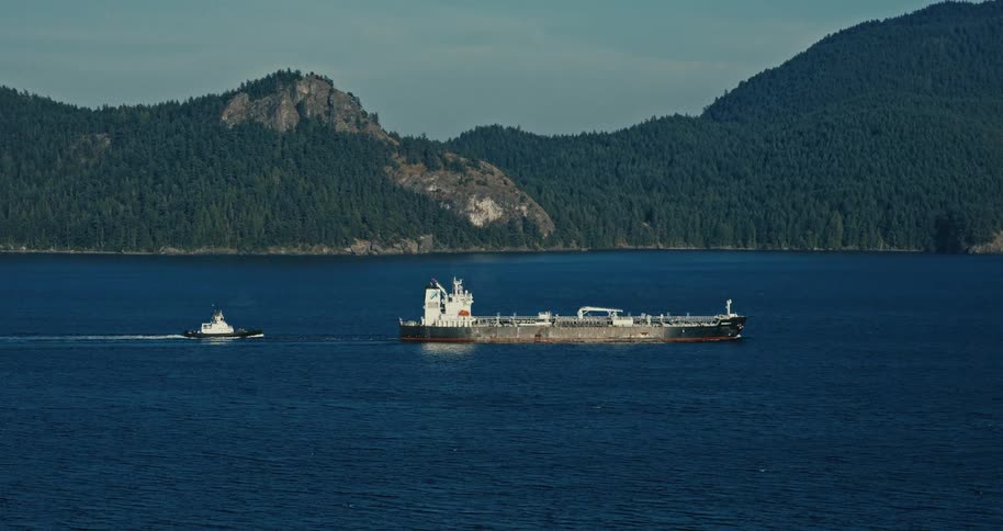 A tugboat tows a large tanker ship through a scenic ocean passage with forested mountains in the background
