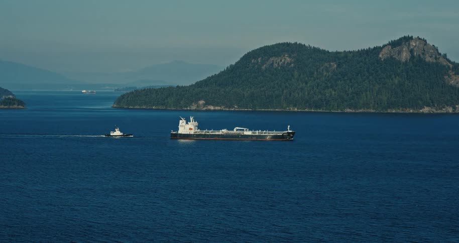A large tanker ship and tugboat navigate a serene blue ocean channel next to a tree-covered island