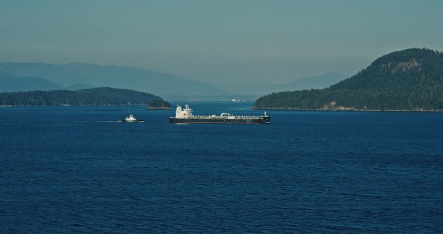 A massive tanker ship accompanied by a tugboat sails through a serene blue channel surrounded by forested islands and distant mountains