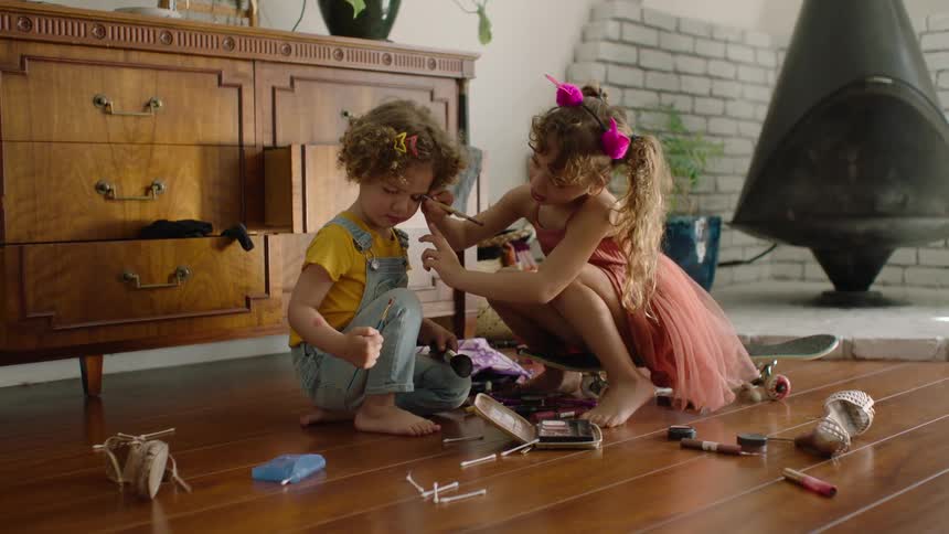Two young sisters play dress-up with makeup on the wooden floor of their sunlit living room