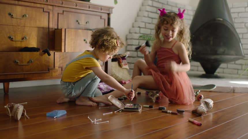 Two cute young sisters play with makeup on the hardwood floor of their sunlit living room
