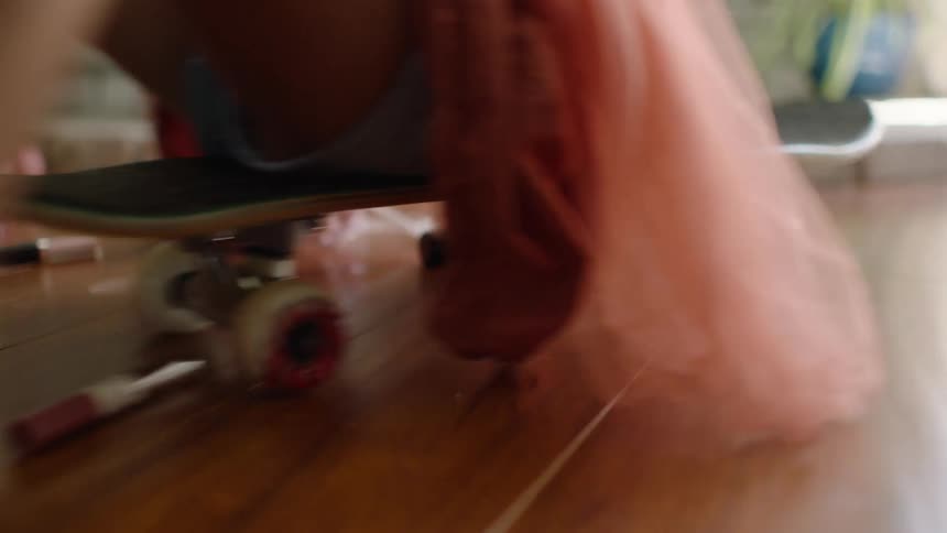 A young child in a pink tutu playfully pushes off on a skateboard, rolling across a hardwood floor past scattered makeup