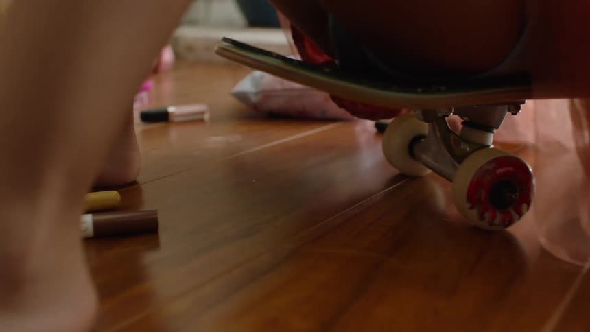 A child on a skateboard rolls through a pile of makeup scattered on a hardwood floor