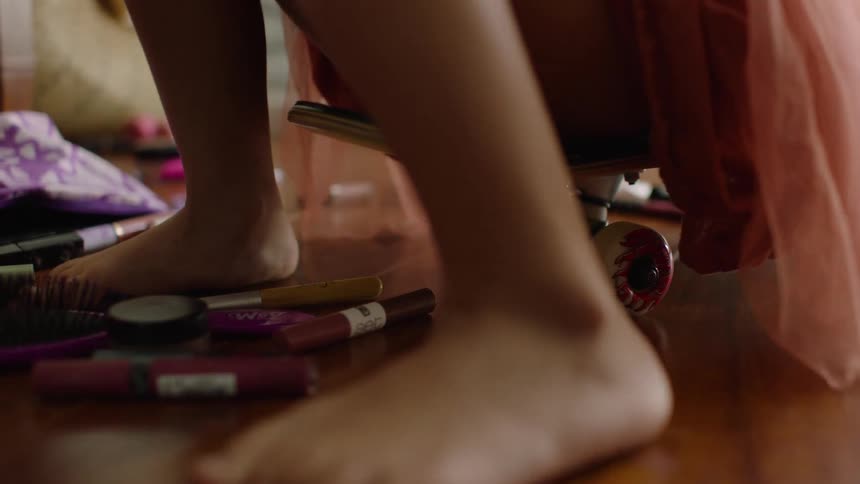 A close-up shot of a young girl sitting on a skateboard and playing with makeup scattered across a wooden floor