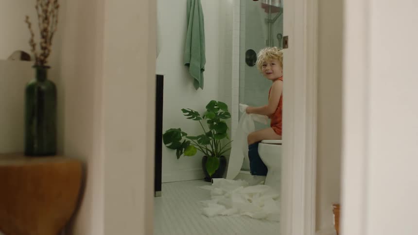 A mischievous little boy laughs while unrolling toilet paper all over the bathroom floor