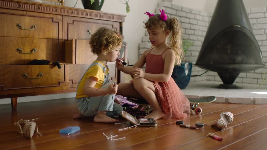 A young girl playfully applies lipstick to her younger brother as they explore a makeup kit on the living room floor