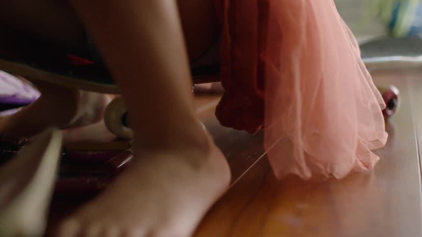 A close-up shot of a young girl's feet as she sits on a skateboard and plays with makeup scattered on the floor