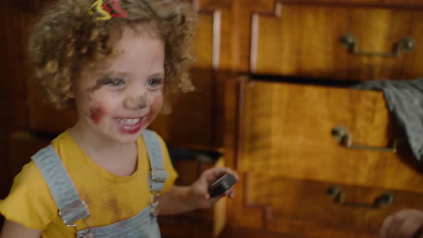 Slow motion close-up of a mischievous little girl with messy makeup on her face smiling while playing with cosmetics