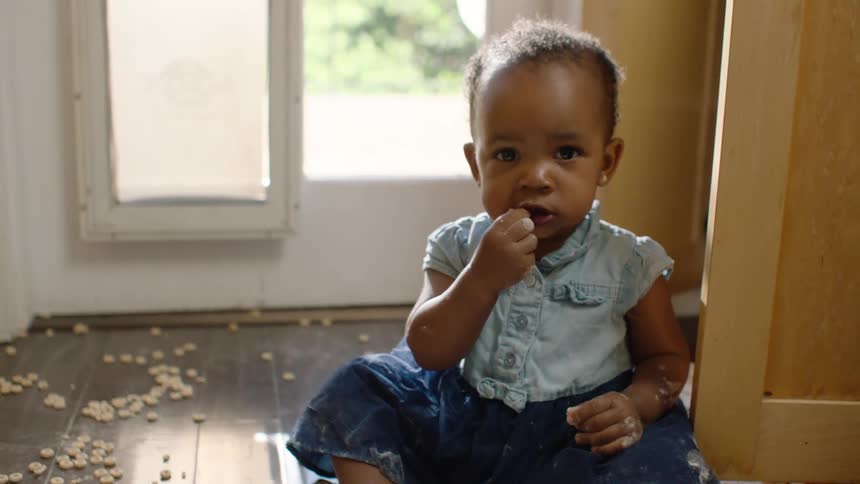 A slow pan reveals an adorable baby girl sitting on the floor in a mess of spilled cereal and flour, happily eating and pointing