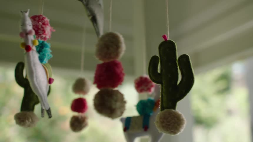 A slow-motion close-up of a whimsical, handmade baby mobile featuring felt cacti, llamas, and colorful pom-poms gently moving