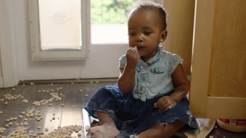 An adorable baby girl sits on the kitchen floor covered in flour, happily eating cereal from a pile she spilled