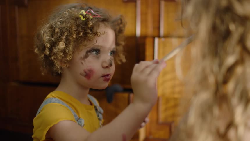 A little girl with curly hair and messy makeup on her face is applying eyeshadow to her sister