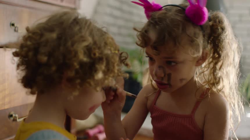 A young girl with messy makeup and bunny ears playfully applies eyeshadow to her brother's face at home