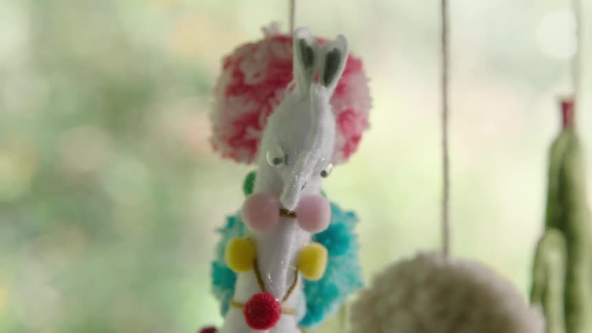 A close-up of a whimsical handmade llama ornament with colorful pom-poms sways gently, eventually drifting out of focus against a soft green background