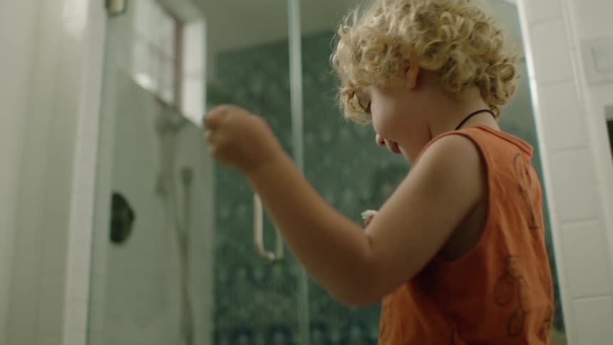A young boy with curly blonde hair learns how to floss his teeth in the bathroom