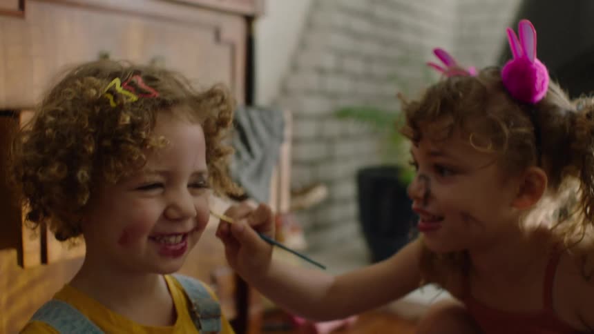 A little girl with curly hair smiles while her sister playfully applies makeup to her face with a small brush