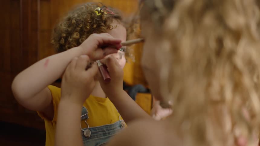 Two adorable little sisters with curly hair have fun playing with their mother's makeup, messily applying it to their faces