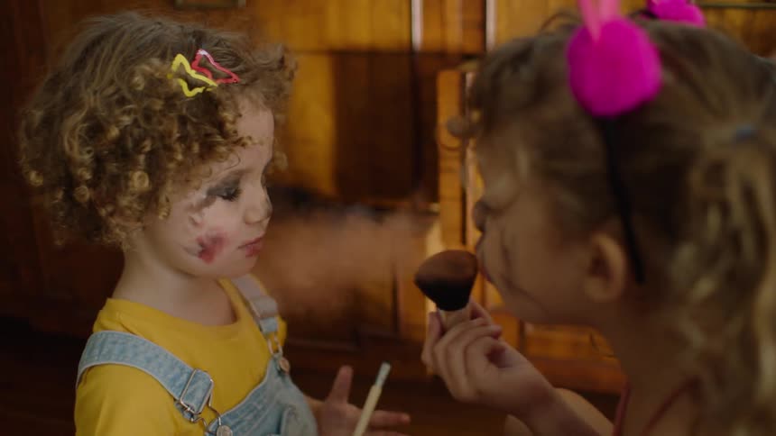 Two young sisters with messy makeup playfully apply powder to each other's faces in slow motion