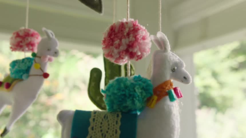 A whimsical handmade baby mobile with colorful felt llamas, cacti, and yarn pom-poms gently sways in a brightly lit nursery