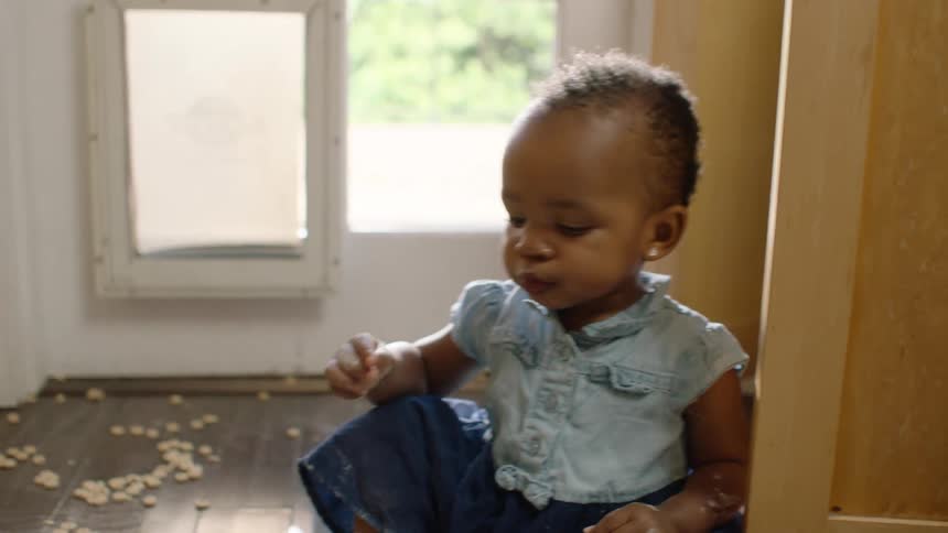 An adorable toddler sits on a wooden floor, making a mess and snacking on spilled cereal