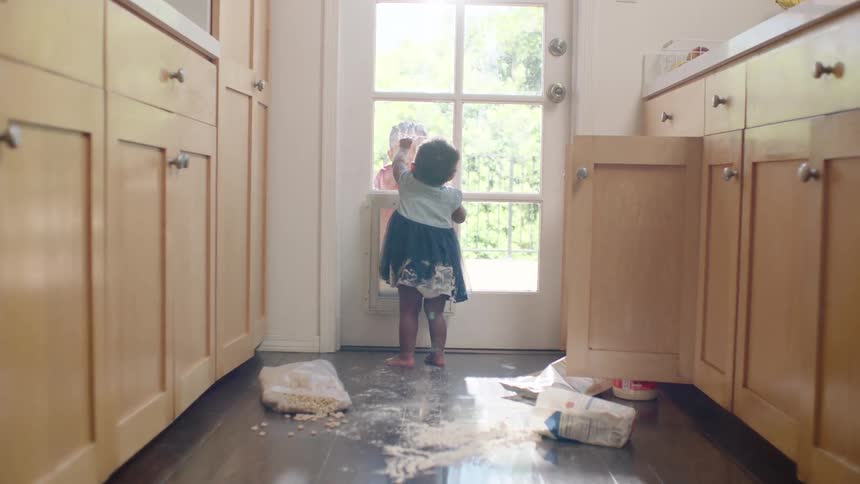 After making a mess in the kitchen, a little girl is greeted by her sibling through the glass back door