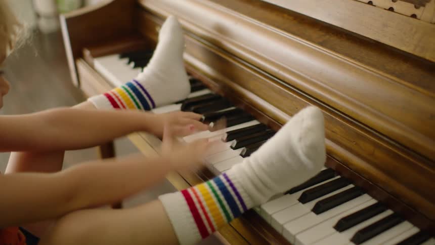 A playful toddler in rainbow socks has fun using their hands and feet to press the keys of an upright piano