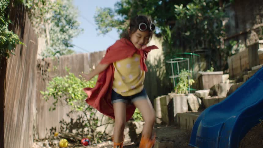 A young superhero girl in a red cape and rain boots joyfully jumps and splashes in a backyard mud puddle in slow motion
