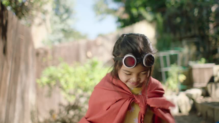 An imaginative young girl with a pirate flag and red cape spins and smiles while playing in her backyard in slow motion