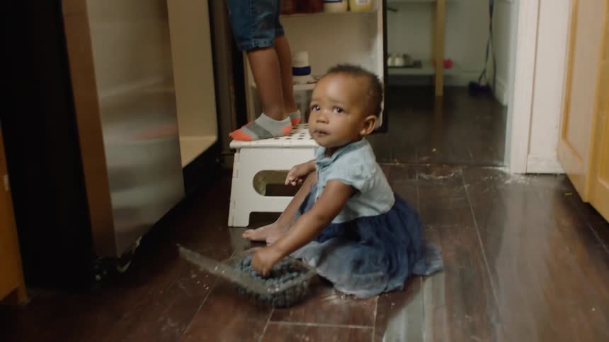 A baby girl eats spilled blueberries on the kitchen floor while her brother gets a drink from the refrigerator