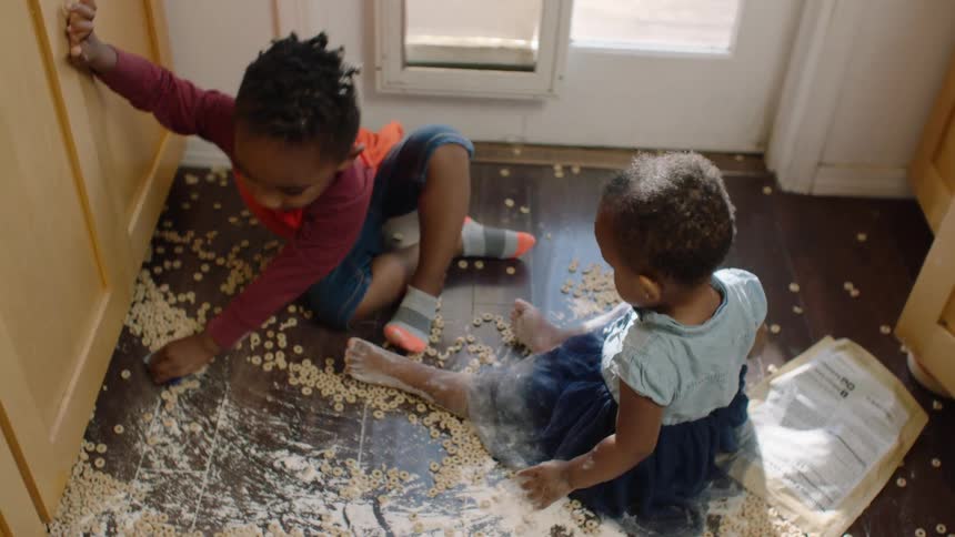 A high-angle view shows a brother and sister playing in a massive spill of cereal and flour on the kitchen floor