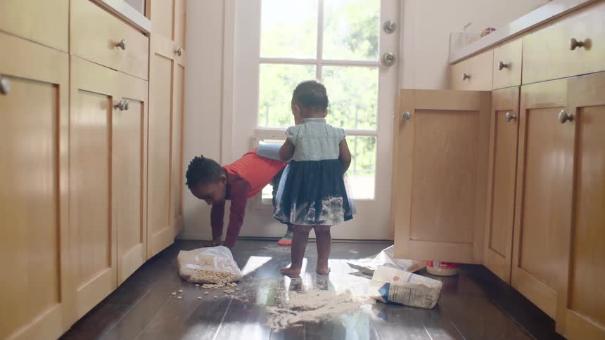 Mischievous toddler siblings play in spilled flour and cereal in a sunlit kitchen