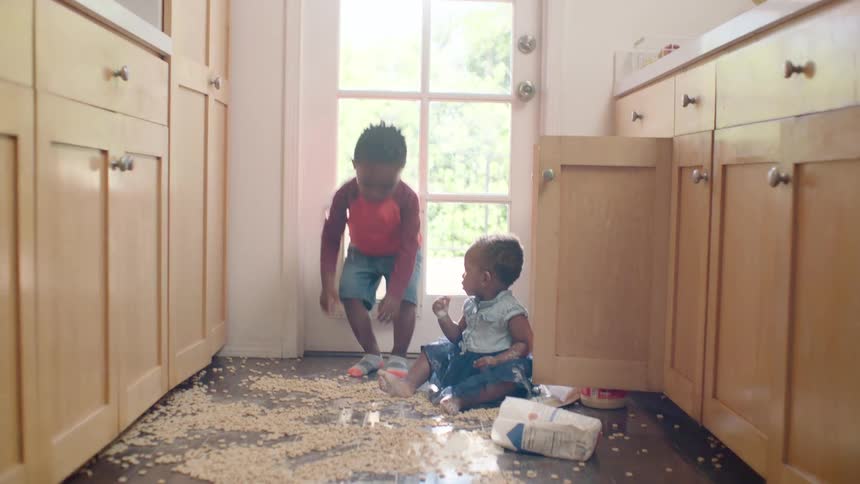 Two young siblings happily play in a huge mess they made with cereal and flour on the kitchen floor