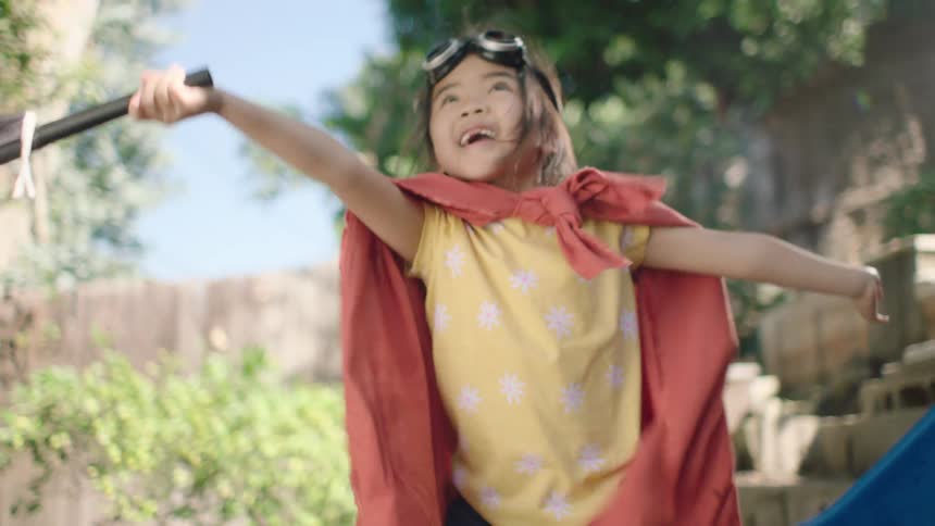 A joyful young girl in a superhero cape and goggles plays pirate, happily jumping in a large mud puddle in her sunny backyard