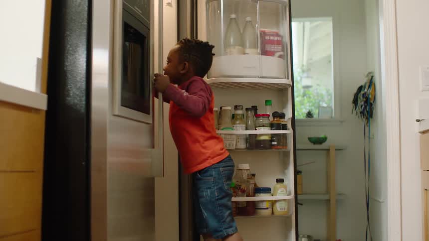 A toddler girl sits on the kitchen floor with blueberries as her brother reaches into the open fridge