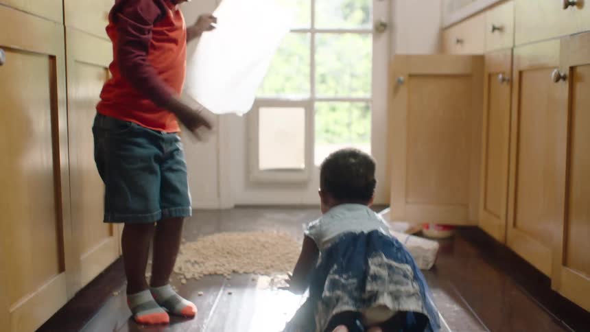 Two young siblings make a huge mess together with cereal and flour on the kitchen floor