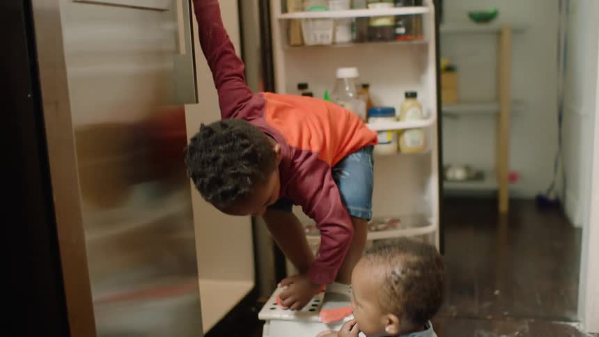 An adorable baby girl eats blueberries on the kitchen floor while her older brother gets a snack from the refrigerator
