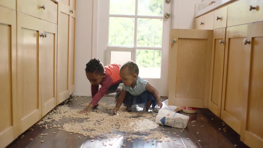 Two mischievous young siblings play together in a huge mess of spilled cereal and flour on the kitchen floor