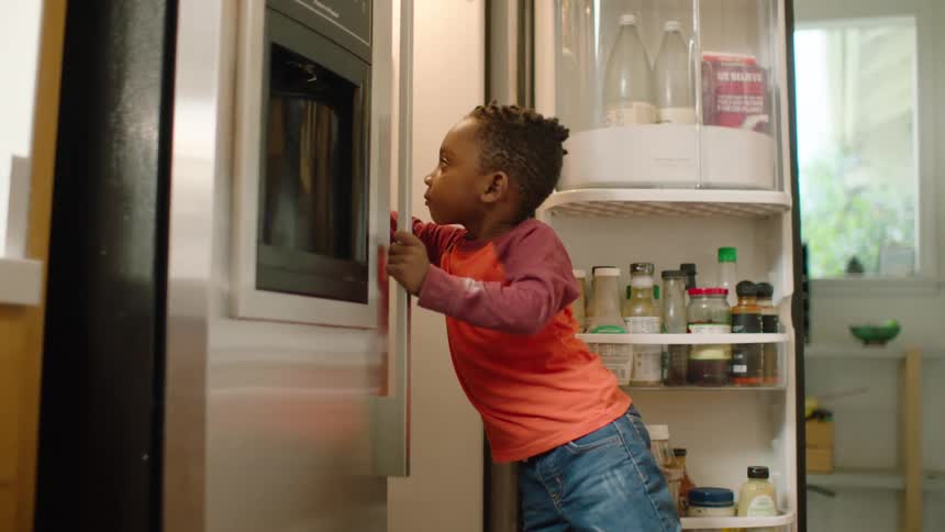 A toddler boy struggles to close the refrigerator door while his baby sister sits on the floor eating blueberries