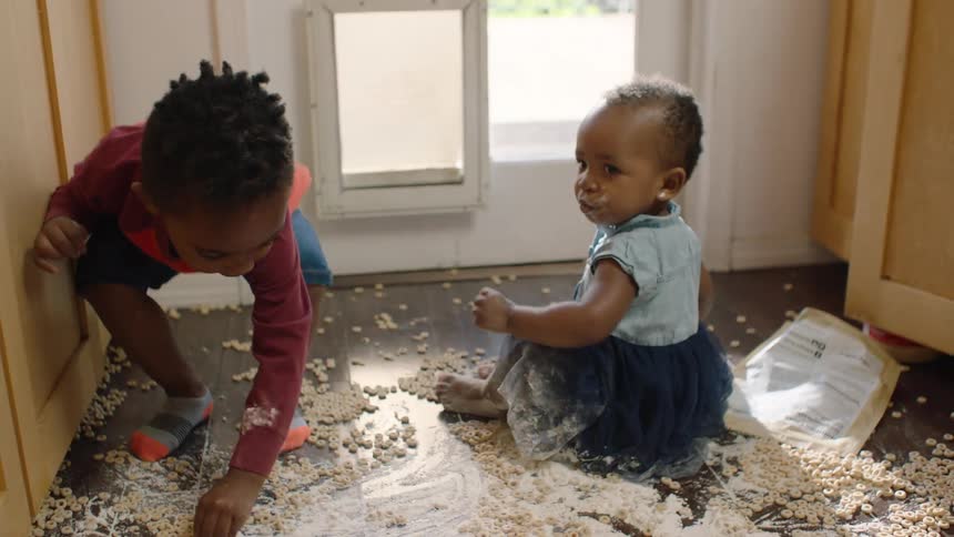 Slow motion shot of a toddler and baby playing together in a huge mess of spilled cereal and flour on the kitchen floor