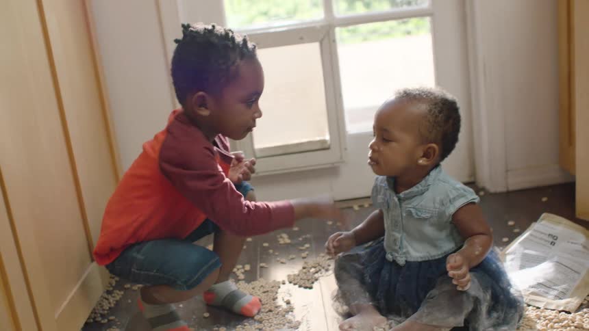 An older brother playfully puts flour on his baby sister's face as they sit in a mess of cereal on the kitchen floor