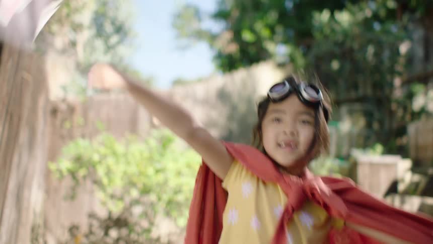 A happy young girl dressed in a red cape and goggles imagines she's a pirate, playfully waving a skull and crossbones flag in her backyard