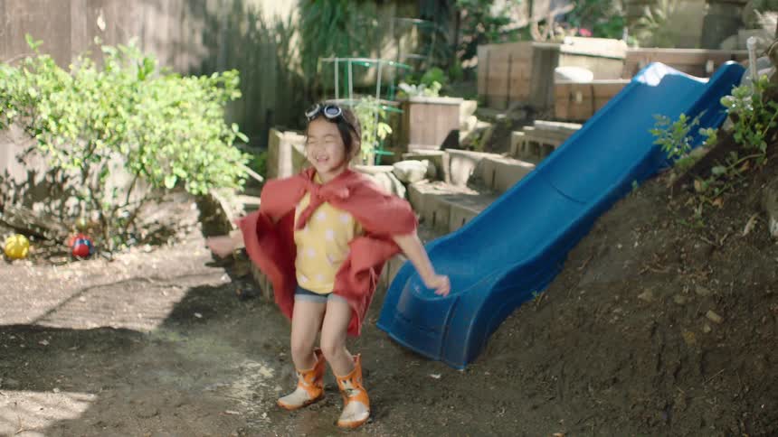 A joyful young girl in a superhero costume with a red cape and goggles jumps and dances in a muddy puddle in her backyard in slow motion